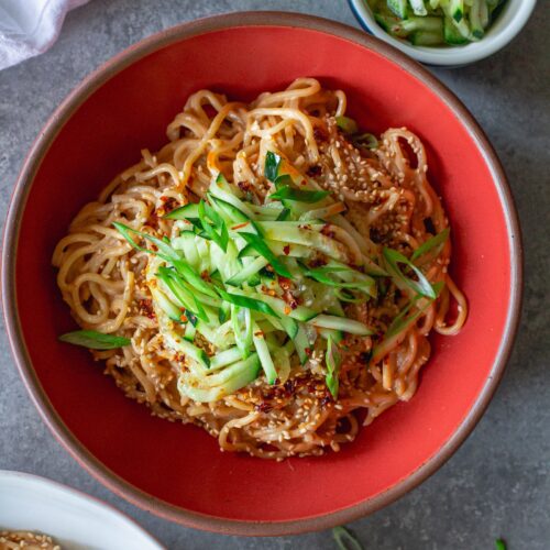 Overhead shot of cold sesame noodles in a bowl topped with sliced cucumbers and toasted sesame seeds.