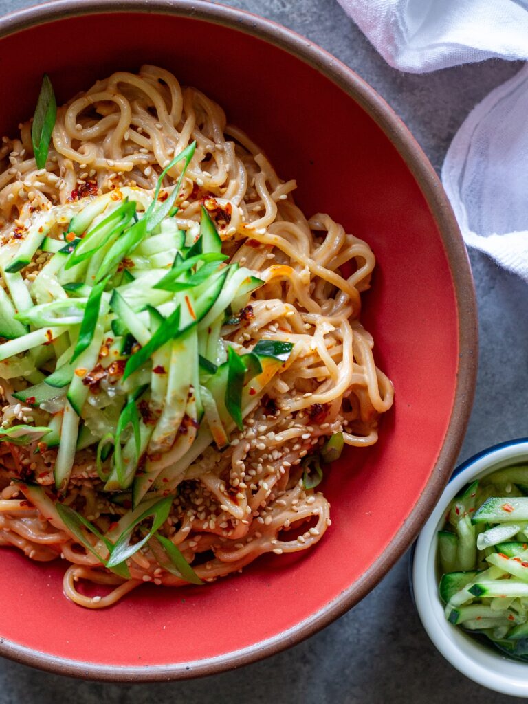 Overhead shot of cold sesame noodles in a bowl topped with sliced cucumbers and toasted sesame seeds.