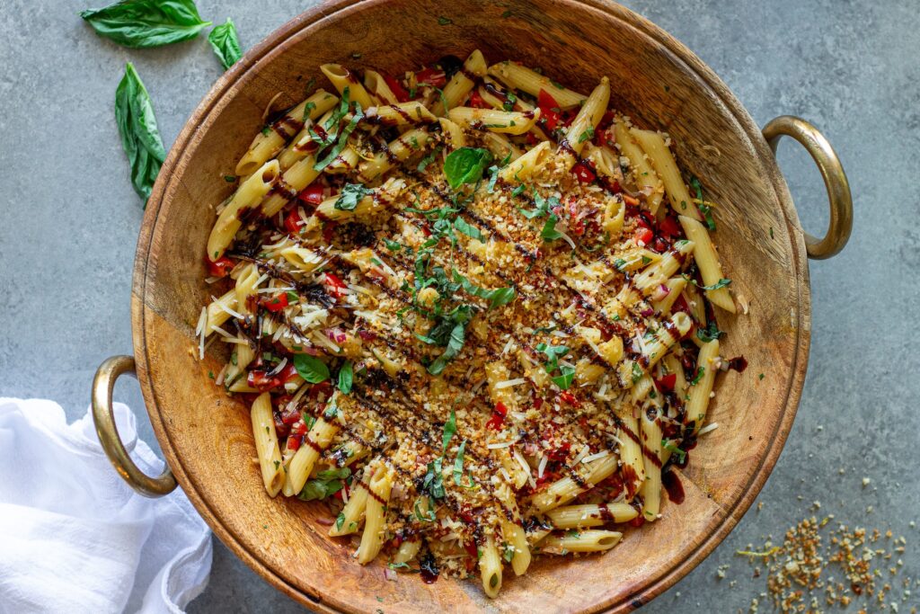 Overhead shot of bruschetta-inspired pasta salad in a serving bowl drizzled with balsamic glaze and garnished with basil and garlicky breadcrumbs.