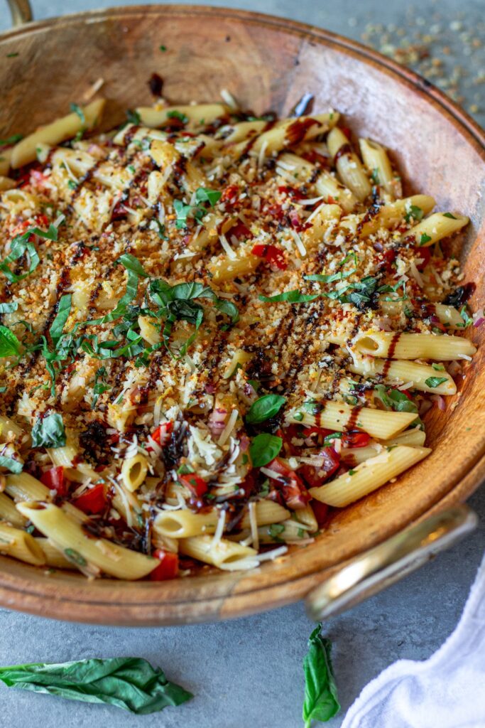 Close-up shot of bruschetta-inspired pasta salad in a serving bowl drizzled with balsamic glaze and garnished with basil and garlicky breadcrumbs.