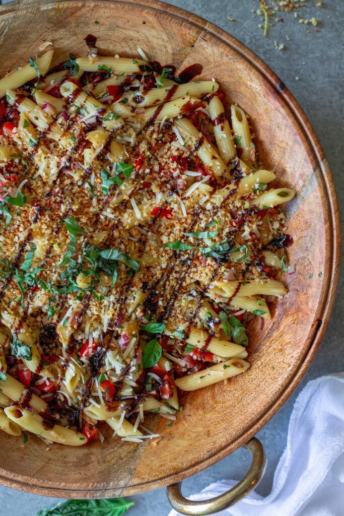 Close-up shot of bruschetta-inspired pasta salad in a serving bowl drizzled with balsamic glaze and garnished with basil and garlicky breadcrumbs.