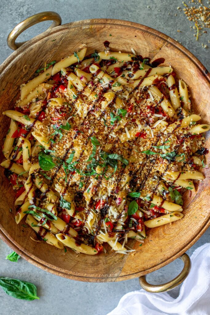 Overhead shot of bruschetta-inspired pasta salad in a serving bowl drizzled with balsamic glaze and garnished with basil and garlicky breadcrumbs.