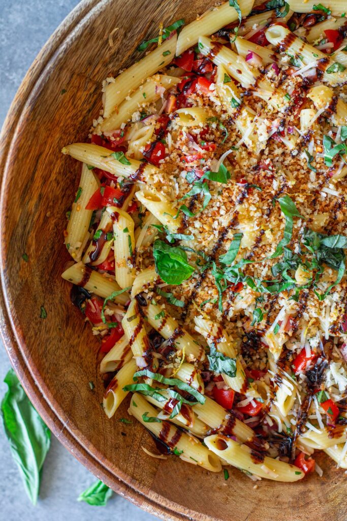 Close-up shot of bruschetta-inspired pasta salad in a serving bowl drizzled with balsamic glaze and garnished with basil and garlicky breadcrumbs.