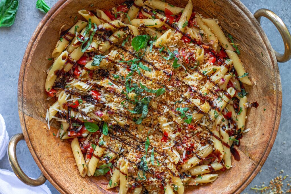 Overhead shot of bruschetta-inspired pasta salad in a serving bowl drizzled with balsamic glaze and garnished with basil and garlicky breadcrumbs.