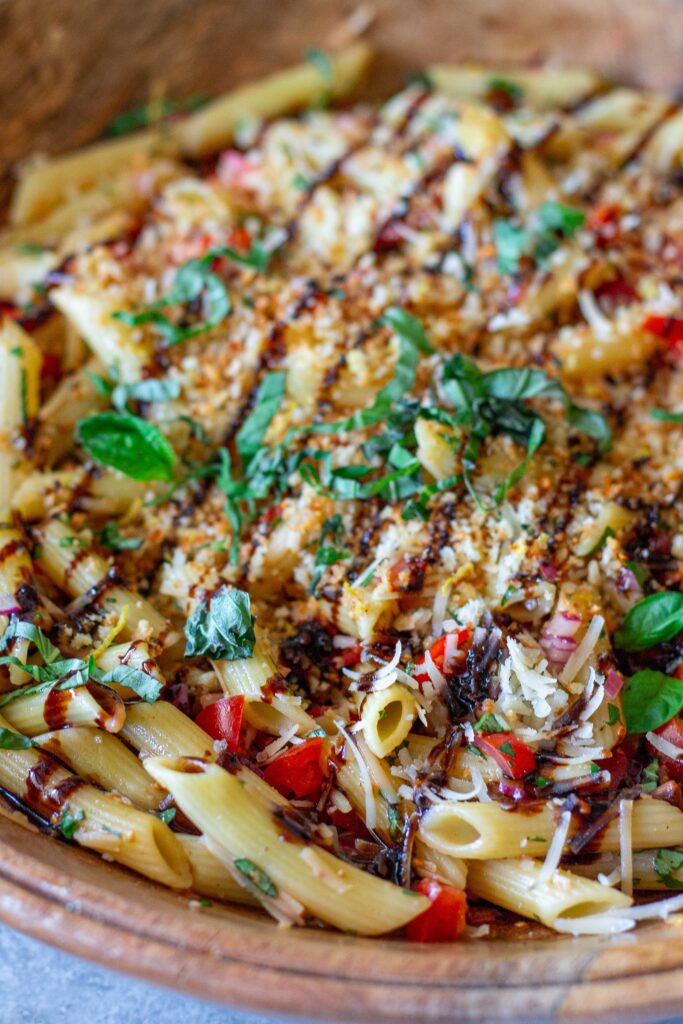 Close-up shot of bruschetta-inspired pasta salad in a serving bowl drizzled with balsamic glaze and garnished with basil and garlicky breadcrumbs.