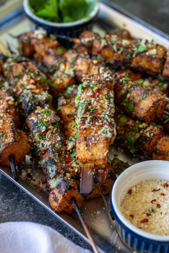 Garlic parmesan tofu skewers brushed with garlic parmesan butter and garnished with parsley, red chili flakes, and grated parmesan.