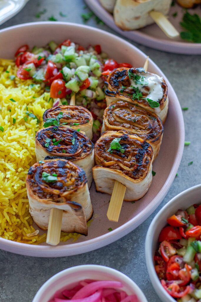 Plated Lula Lavash Roll Kebabs with skewers, creamy garlic drizzle, fresh herbs, turmeric rice, and a refreshing tomato cucumber side salad.