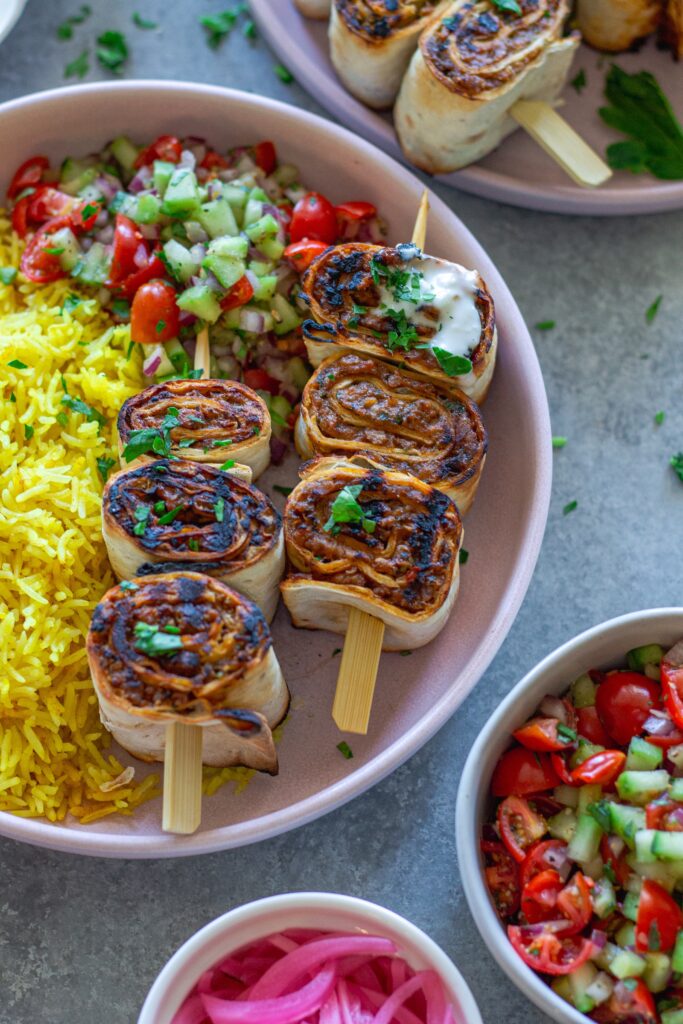 Plated Lula Lavash Roll Kebabs with skewers, creamy garlic drizzle, fresh herbs, turmeric rice, and a refreshing tomato cucumber side salad.