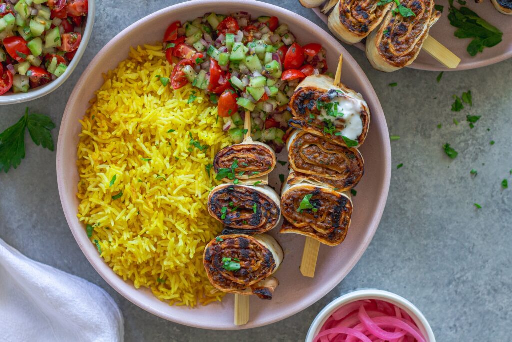 Plated Lula Lavash Roll Kebabs with skewers, creamy garlic drizzle, fresh herbs, turmeric rice, and a refreshing tomato cucumber side salad.