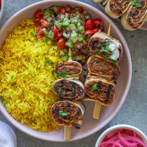 Plated Lula Lavash Roll Kebabs with skewers, creamy garlic drizzle, fresh herbs, turmeric rice, and a refreshing tomato cucumber side salad.