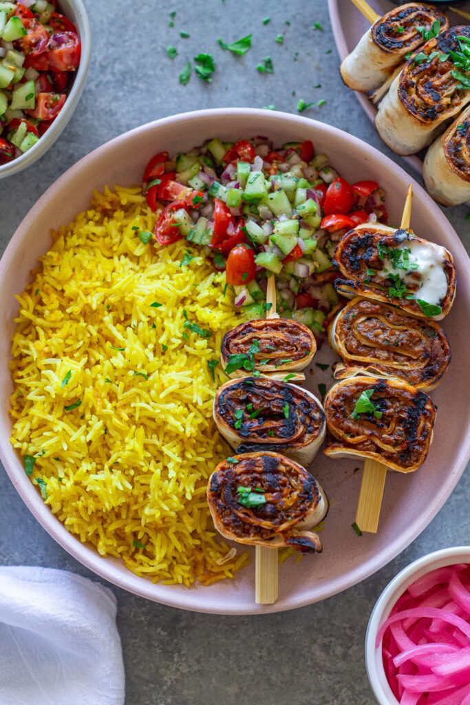 Plated Lula Lavash Roll Kebabs with skewers, creamy garlic drizzle, fresh herbs, turmeric rice, and a refreshing tomato cucumber side salad.