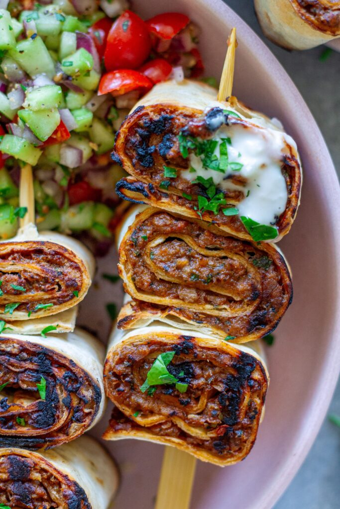 Plated Lula Lavash Roll Kebabs with skewers, creamy garlic drizzle, fresh herbs, turmeric rice, and a refreshing tomato cucumber side salad.