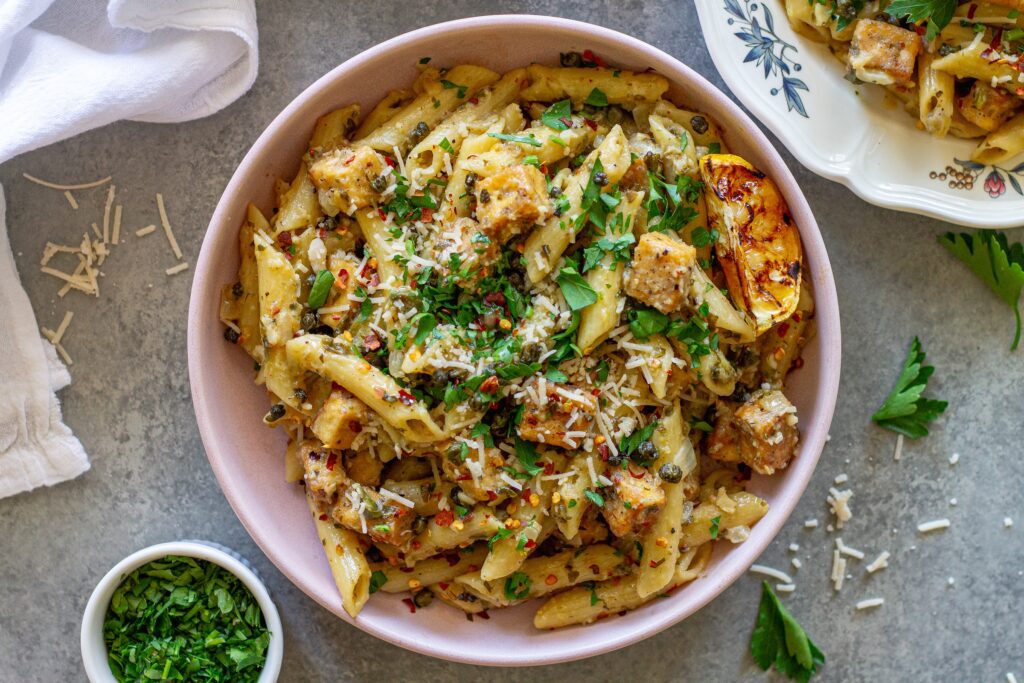 Overhead shot of one pan tofu piccata pasta topped with fresh parsley, fried capers, a sprinkle of parmesan and a grilled lemon slice.