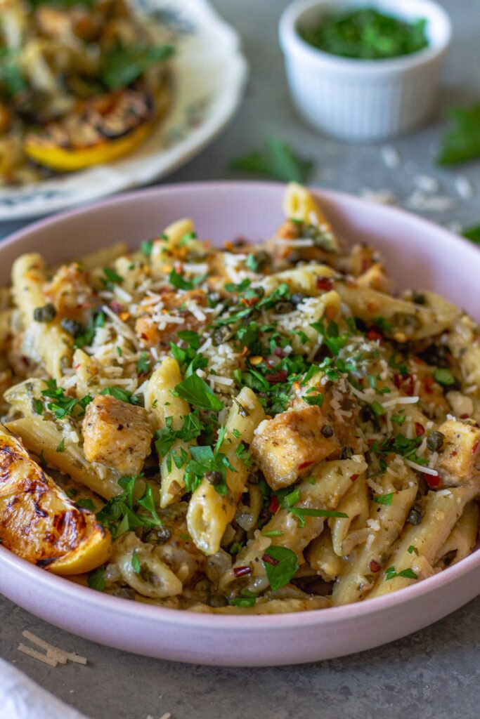 image of one pan tofu piccata pasta topped with fresh parsley, fried capers, a sprinkle of parmesan and a grilled lemon slice.