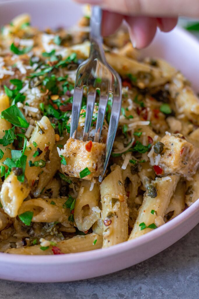a close-up of one pan tofu piccata pasta served in a bowl, with a fork scooping up a bite, topped with vegan parmesan and fresh parsley.