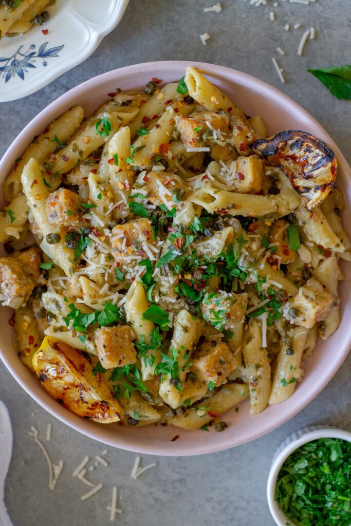 Overhead shot of one pan tofu piccata pasta topped with fresh parsley, fried capers, a sprinkle of parmesan and a grilled lemon slice.