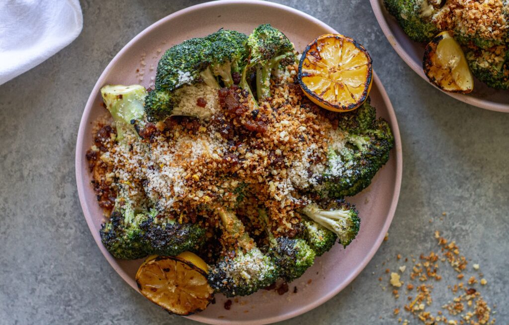 Platter of grilled broccoli Caesar topped with grated parmesan, crispy tofu bacon and lemony breadcrumbs with grilled lemon slices on the side.