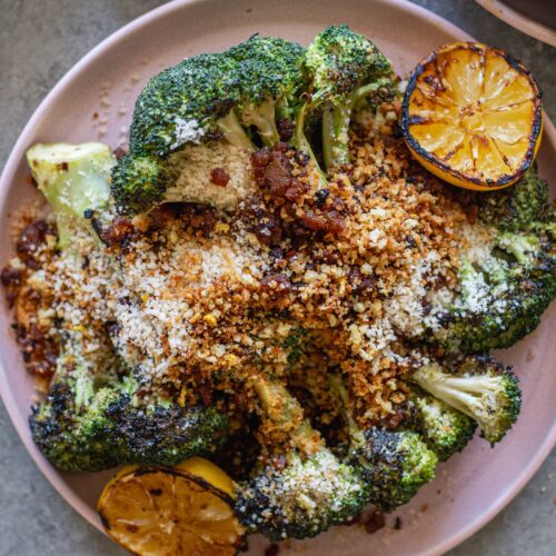 Platter of grilled broccoli Caesar topped with grated parmesan, crispy tofu bacon and lemony breadcrumbs with grilled lemon slices on the side.