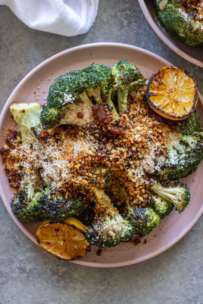 Platter of grilled broccoli Caesar topped with grated parmesan, crispy tofu bacon and lemony breadcrumbs with grilled lemon slices on the side.