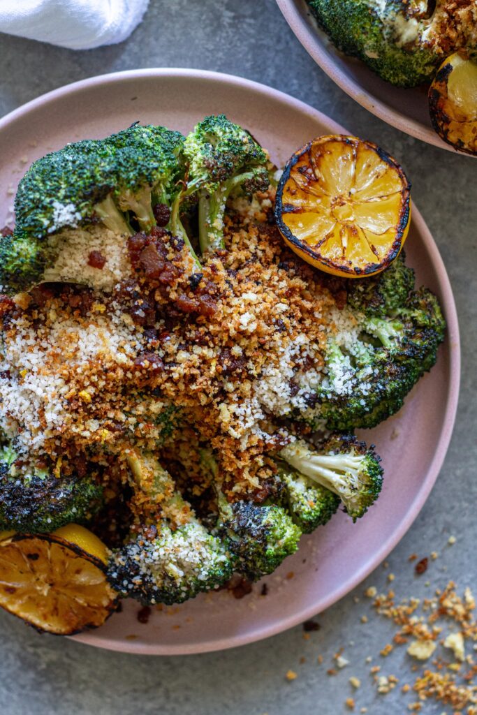 Platter of grilled broccoli Caesar topped with grated parmesan, crispy tofu bacon and lemony breadcrumbs with grilled lemon slices on the side.
