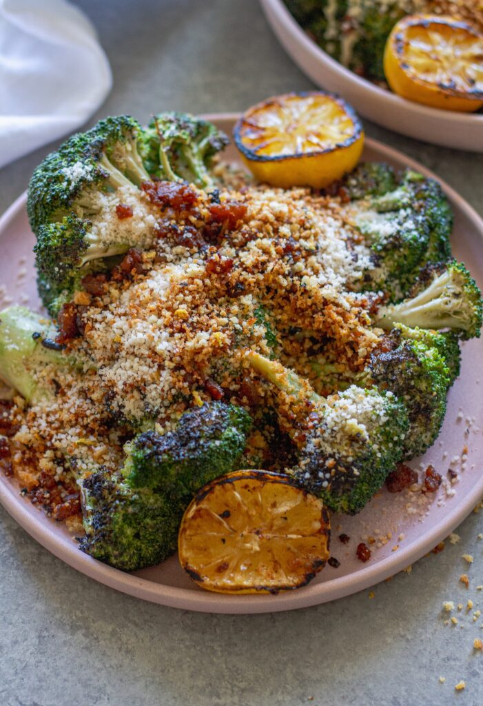 Platter of grilled broccoli Caesar topped with grated parmesan, crispy tofu bacon and lemony breadcrumbs with grilled lemon slices on the side.