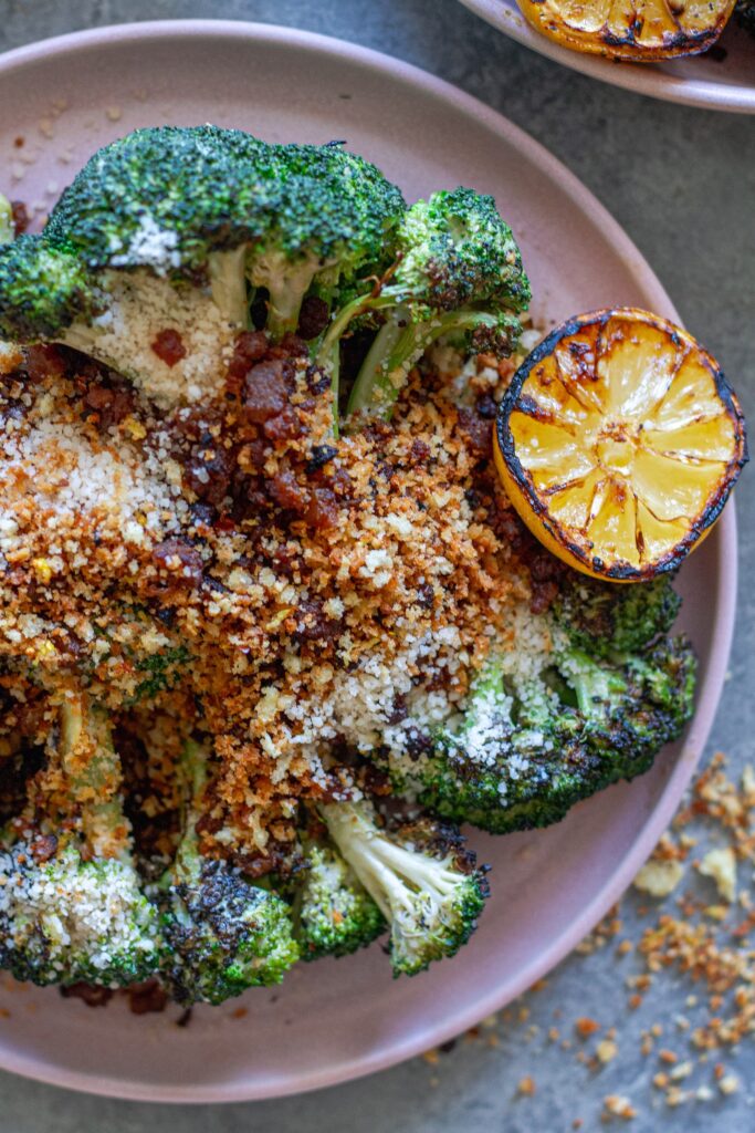 Platter of grilled broccoli Caesar topped with grated parmesan, crispy tofu bacon and lemony breadcrumbs with grilled lemon slices on the side.