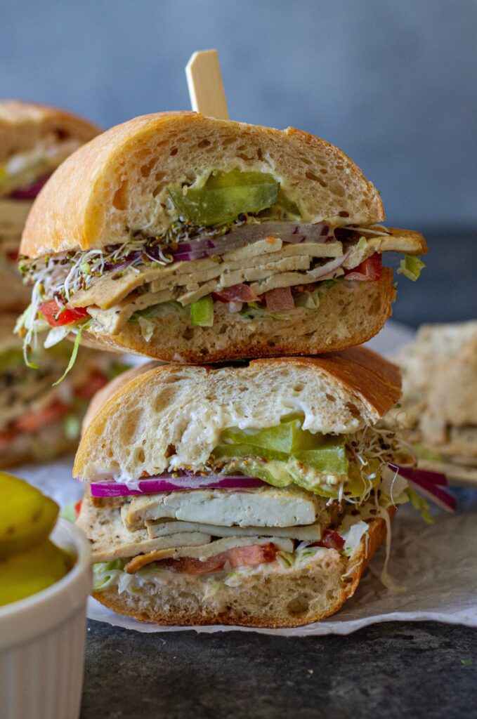 Tofu deli meat slices arranged in a sandwich with pickles, lettuce, tomato, sprouts, and onion.