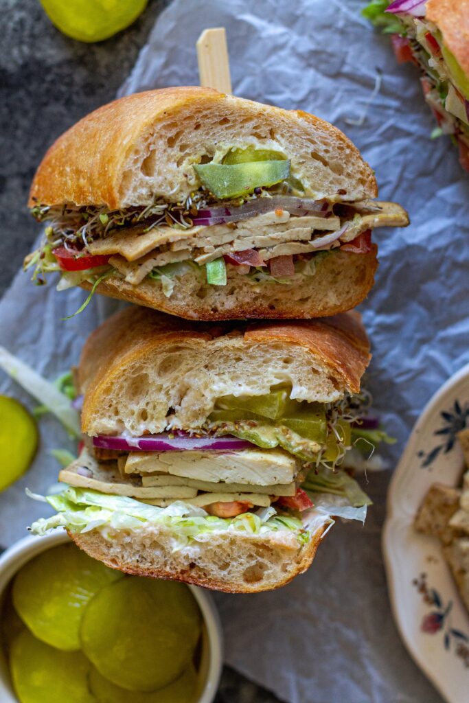 Tofu deli meat slices arranged in a sandwich with pickles, lettuce, tomato, sprouts, and onion.