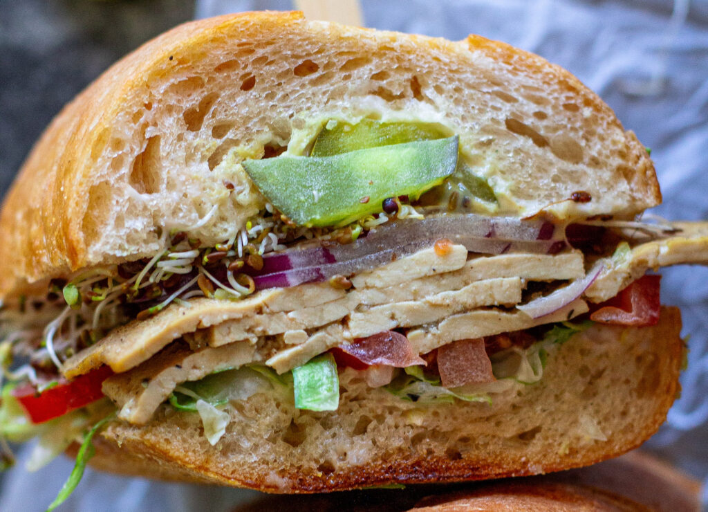 Tofu deli meat slices arranged in a sandwich with pickles, lettuce, tomato, sprouts, and onion.