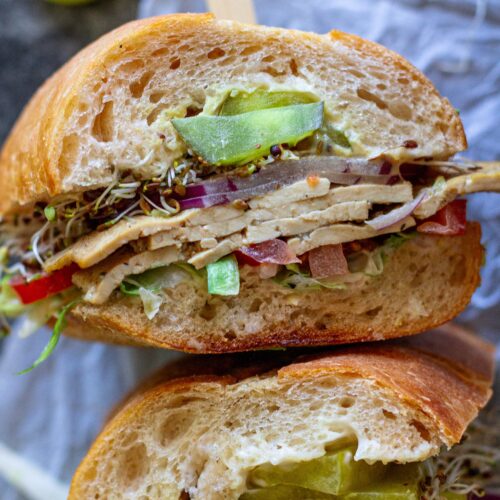Tofu deli meat slices arranged in a sandwich with pickles, lettuce, tomato, sprouts, and onion.