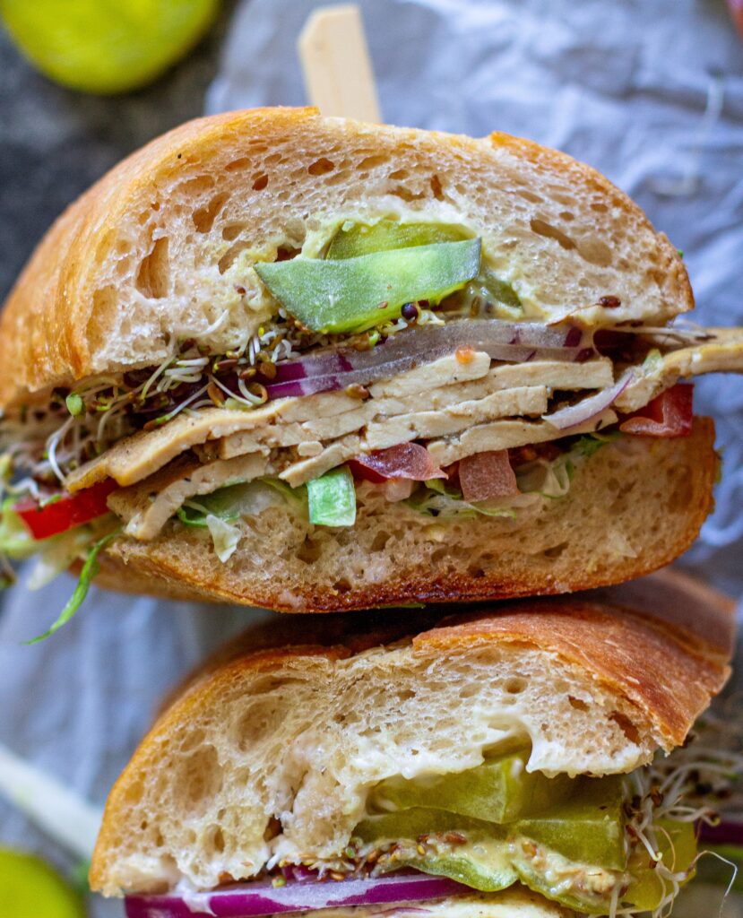 Tofu deli meat slices arranged in a sandwich with pickles, lettuce, tomato, sprouts, and onion.