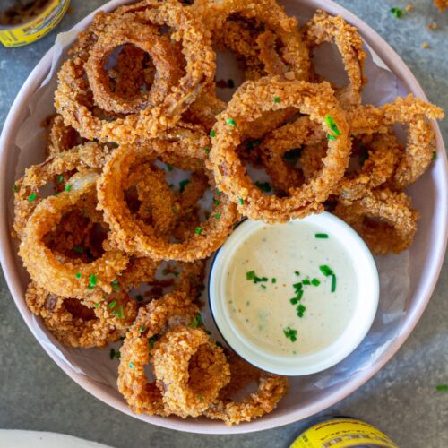 Golden fried onion rings stacked on a plate with a side of white BBQ sauce (aka Alabama sauce) for dipping.