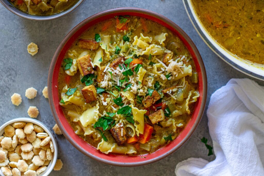Creamy vegan chicken noodle soup garnished with parsley and parmesan, and served with a side of oyster crackers.