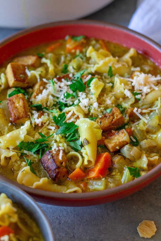 Creamy vegan chicken noodle soup garnished with parsley and parmesan, and served with a side of oyster crackers.