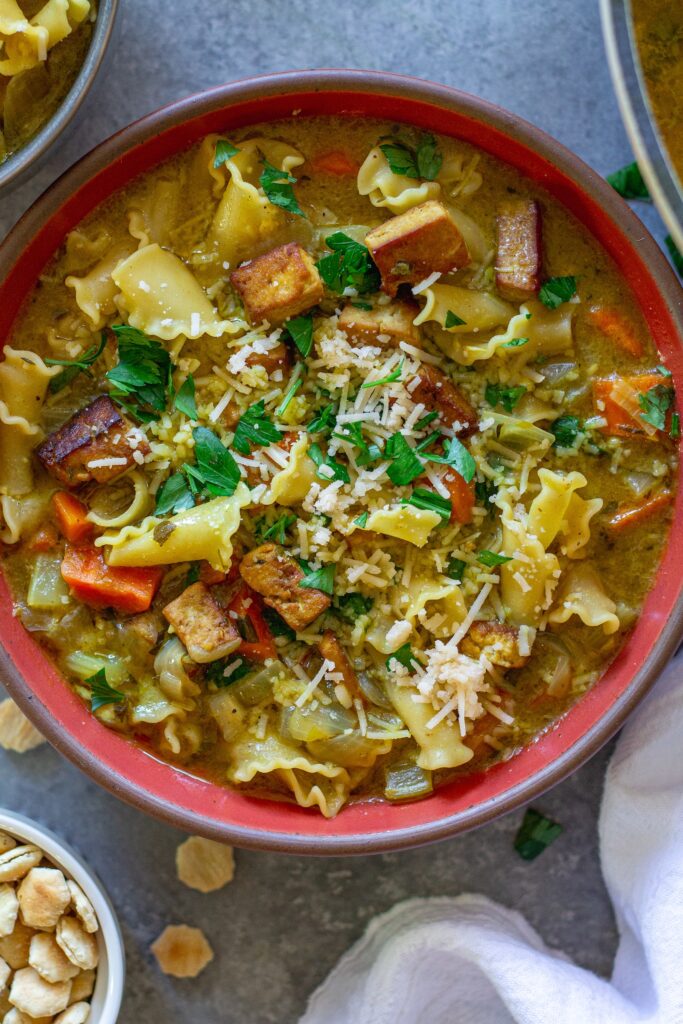 Creamy vegan chicken noodle soup garnished with parsley and parmesan, and served with a side of oyster crackers.