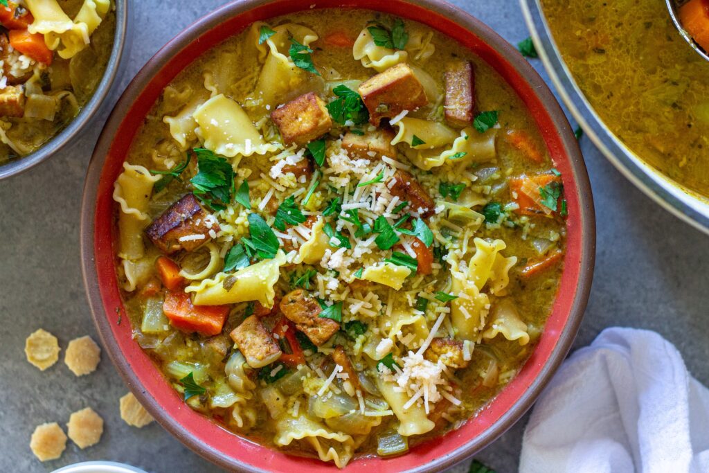Creamy vegan chicken noodle soup garnished with parsley and parmesan, and served with a side of oyster crackers.