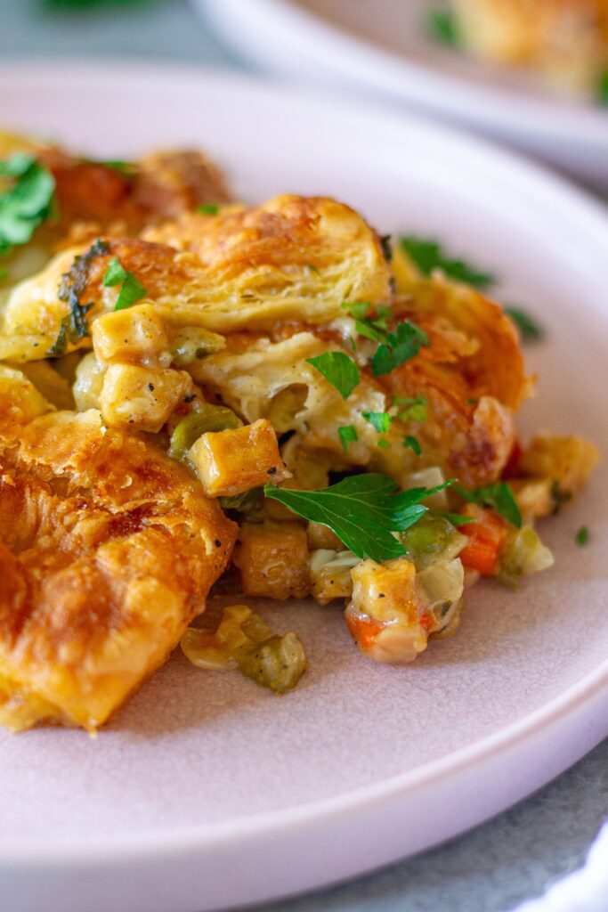 Close up shot of vegan tofu pot pie with creamy veggie filling, topped with golden and flaky puff pastry squares, then garnished with fresh parsley.