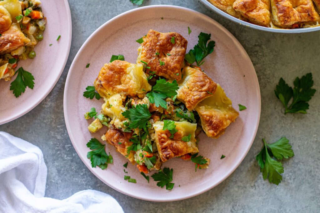 Overhead shot of vegan tofu pot pie with creamy veggie filling, topped with golden and flaky puff pastry squares, then garnished with fresh parsley.