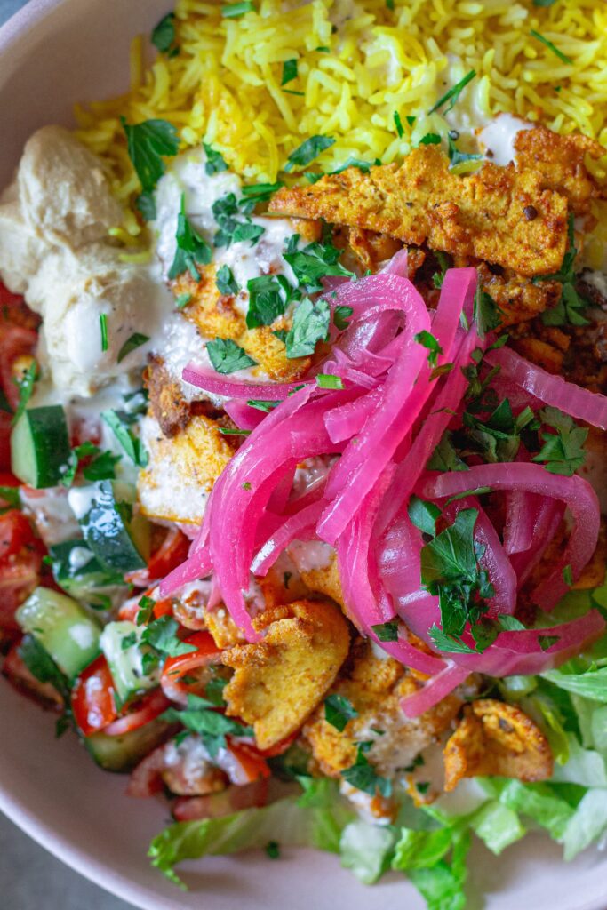 Vibrant tofu shawarma bowl featuring turmeric rice, spiced tofu, cucumber tomato salad, lettuce, and creamy garlic sauce.