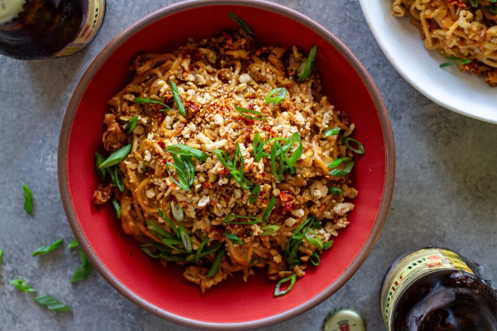 A vibrant bowl of peanut noodle stir fry topped with crispy shredded tofu, caramelized cabbage, green onions, and sesame seeds.