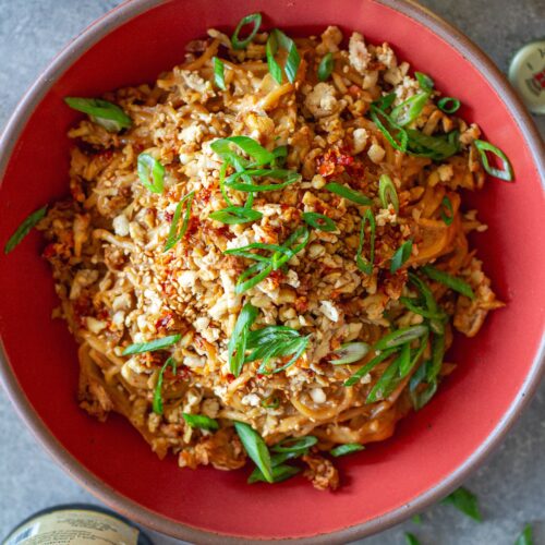 A vibrant bowl of peanut noodle stir fry topped with crispy shredded tofu, caramelized cabbage, green onions, and sesame seeds.