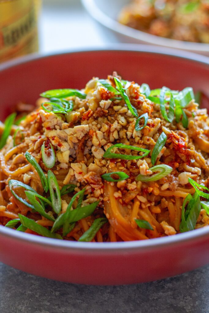 A vibrant bowl of peanut noodle stir fry topped with crispy shredded tofu, caramelized cabbage, green onions, and sesame seeds.