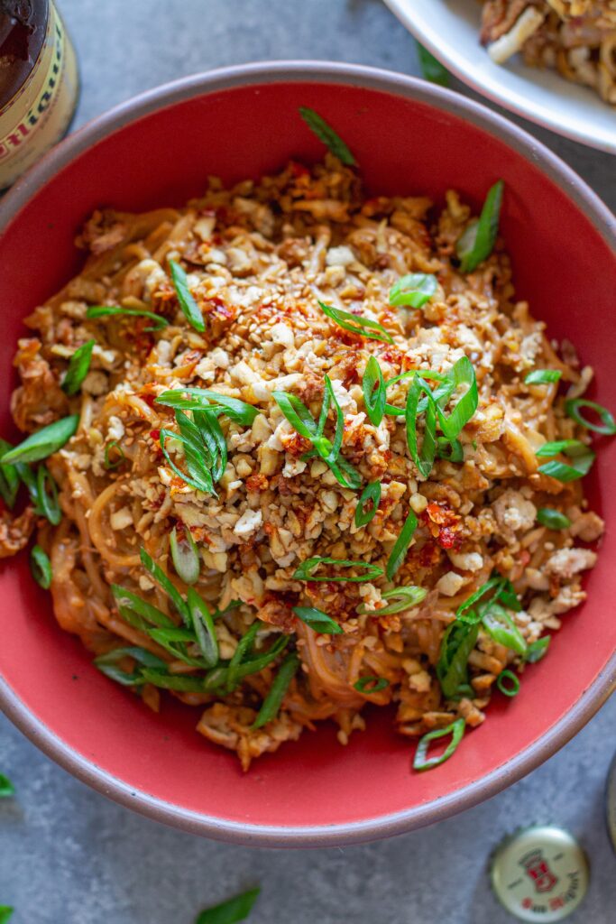 A vibrant bowl of peanut noodle stir fry topped with crispy shredded tofu, caramelized cabbage, green onions, and sesame seeds.