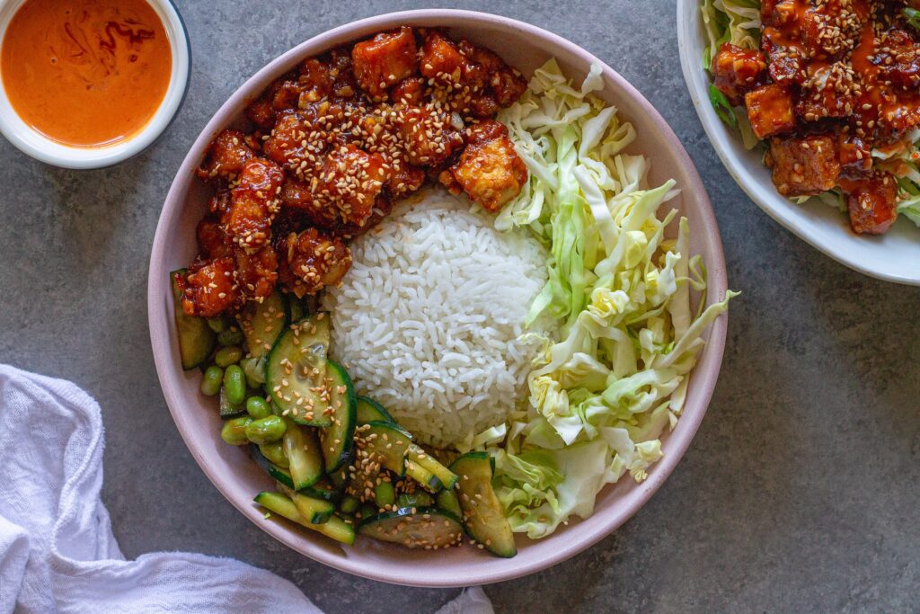 Close‑up of a assembled sticky sriracha tofu bowl with rice, cabbage, edamame, cucumber, and lime wedge.