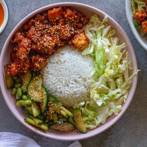 Close‑up of a assembled sticky sriracha tofu bowl with rice, cabbage, edamame, cucumber, and lime wedge.