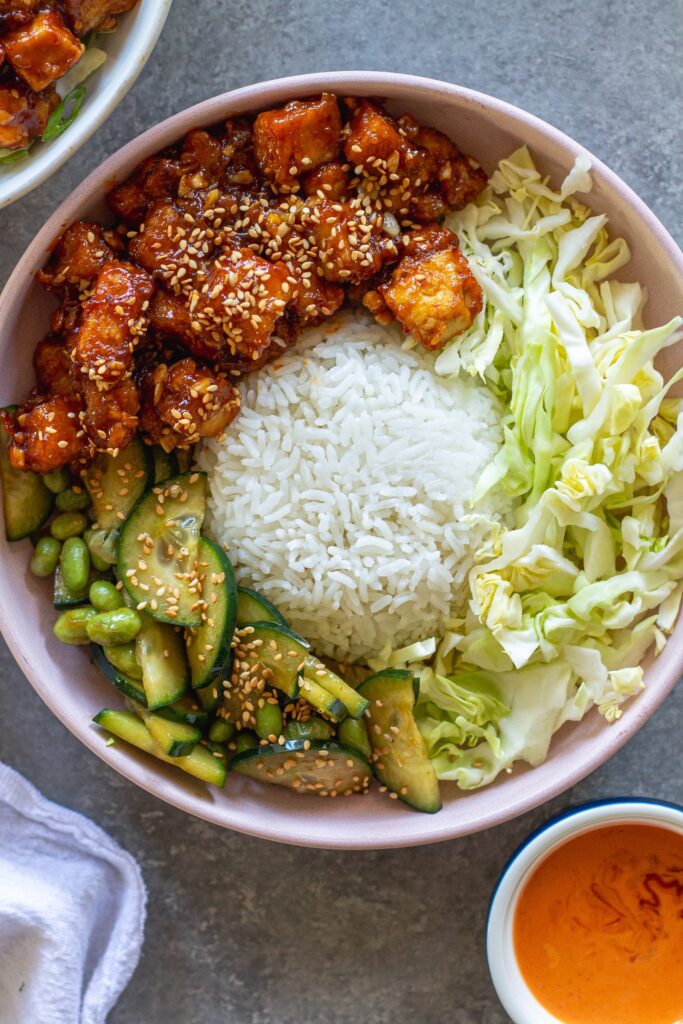 Close‑up of a assembled sticky sriracha tofu bowl with rice, cabbage, edamame, cucumber, and lime wedge.