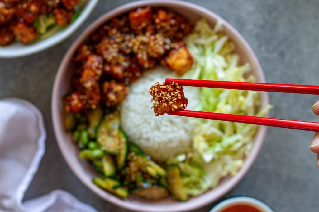 Close‑up of a assembled sticky sriracha tofu bowl with rice, cabbage, edamame, cucumber, and lime wedge.