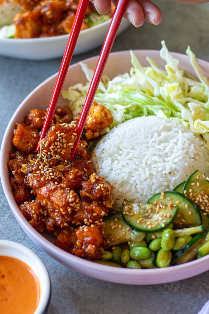 Close‑up of a assembled sticky sriracha tofu bowl with rice, cabbage, edamame, cucumber, and lime wedge.