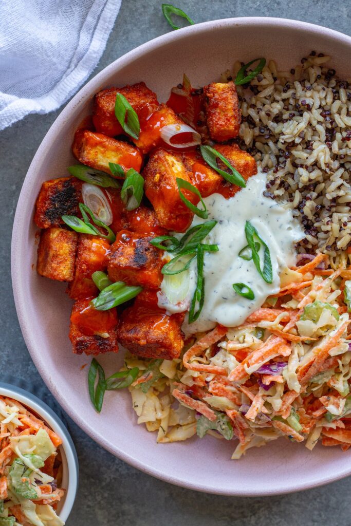 Close-up of a colorful Buffalo Tofu Ranch Bowl with crispy golden tofu, creamy ranch slaw, and quinoa/brown rice mixture, garnished with green onions.