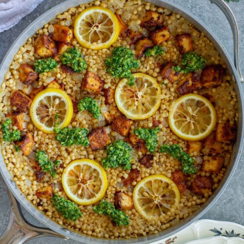 Golden crispy tofu cubes and fluffy lemon couscous in a skillet topped with green chimichurri sauce and lemon slices.